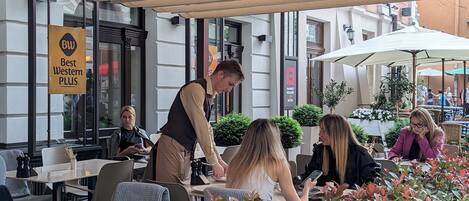An outdoor cafe area with tables and chairs, a waiter attending to a customer, and a sign indicating 'Best Western PLUS' hotel.
