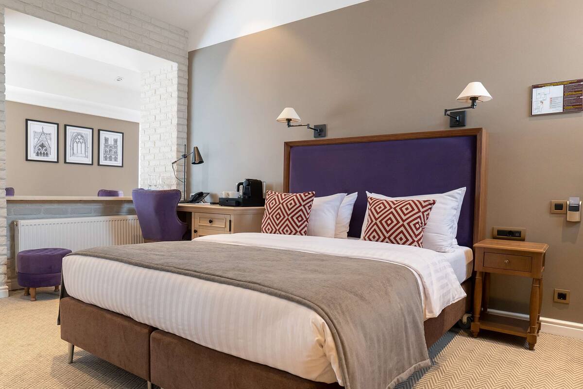 A modern hotel room with a large bed, a desk with a computer, a chair, and a small table with a lamp.