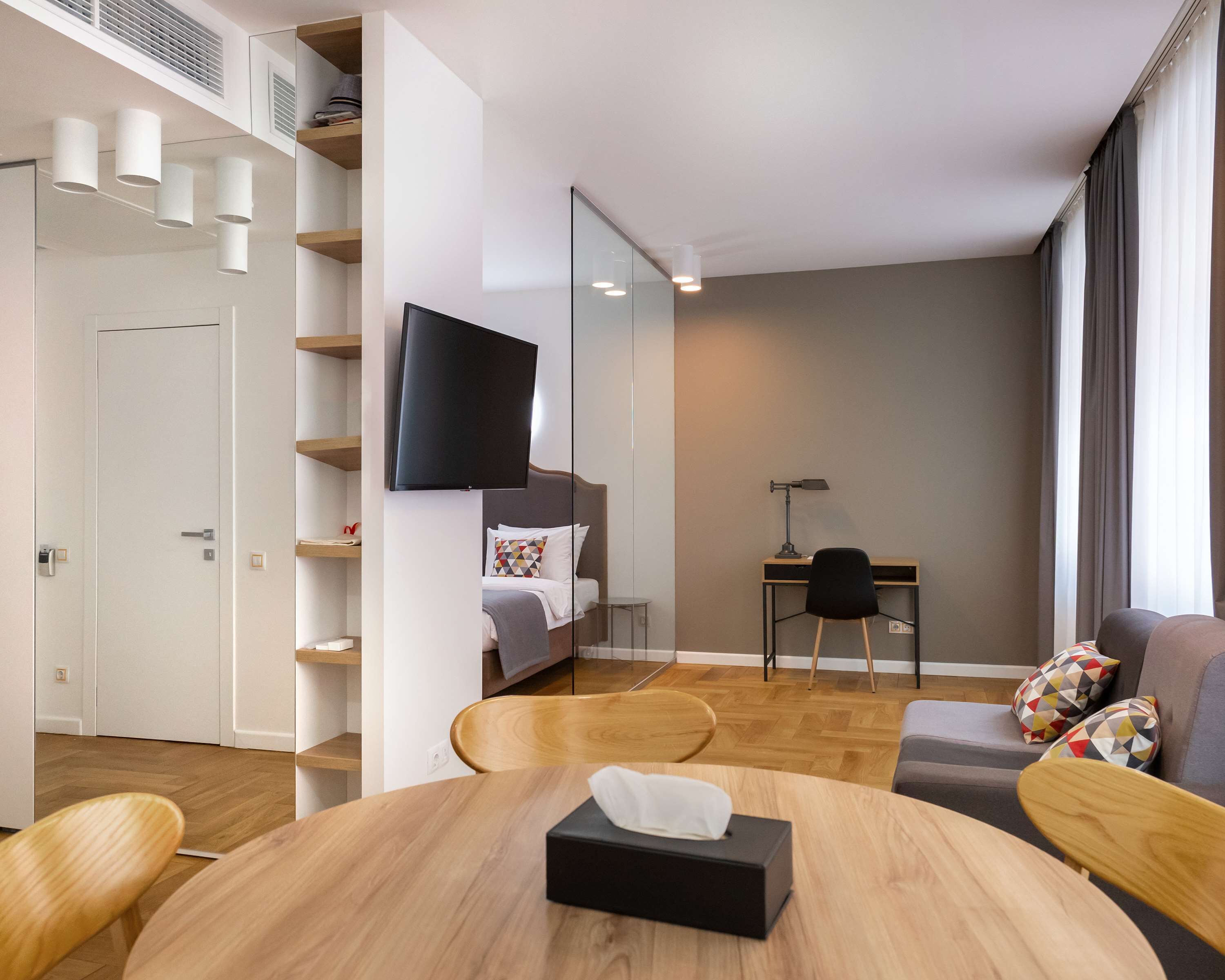 A modern hotel room with a bed, a desk, a TV, and a dining area with a table and chairs.