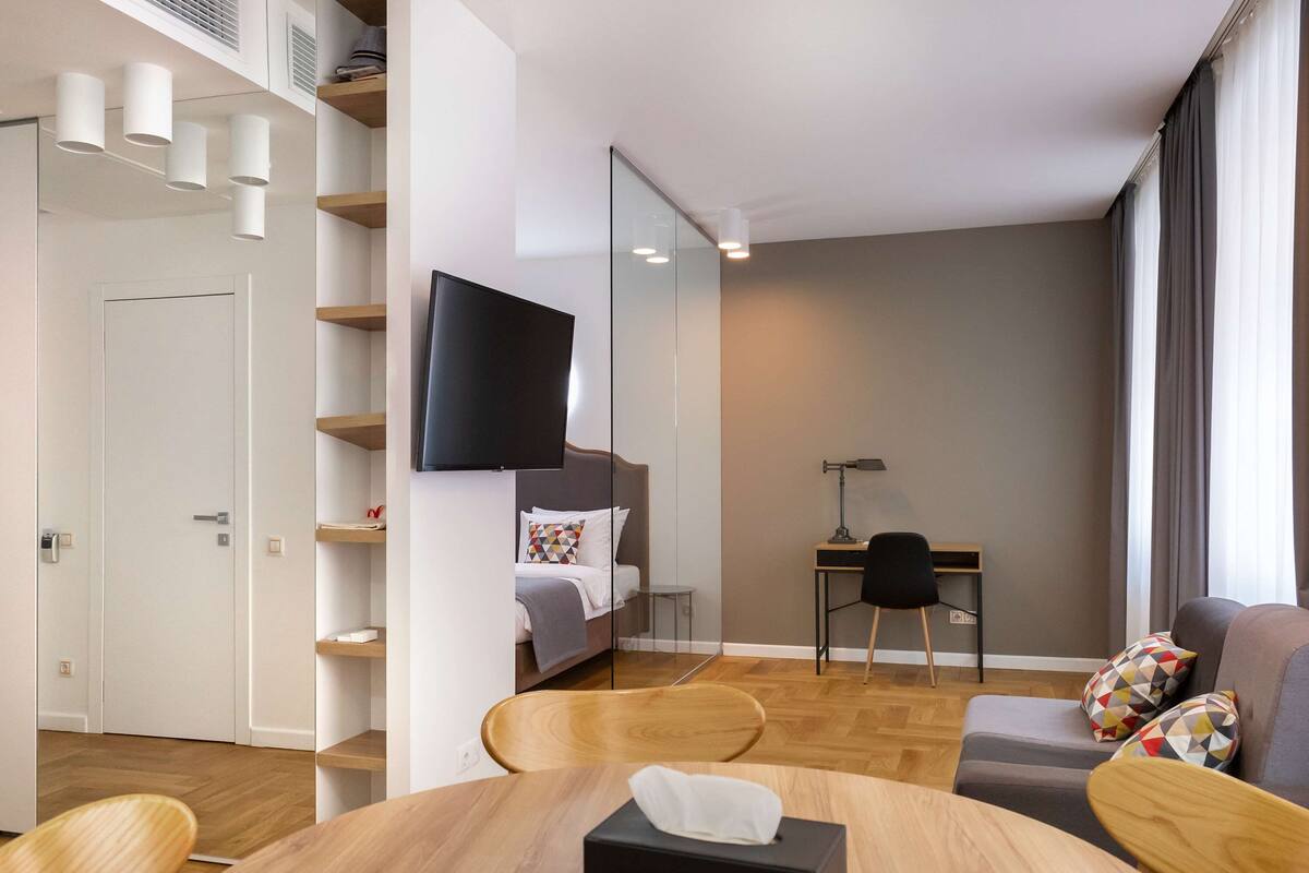 A modern hotel room with a bed, a desk, a TV, and a dining area with a table and chairs.