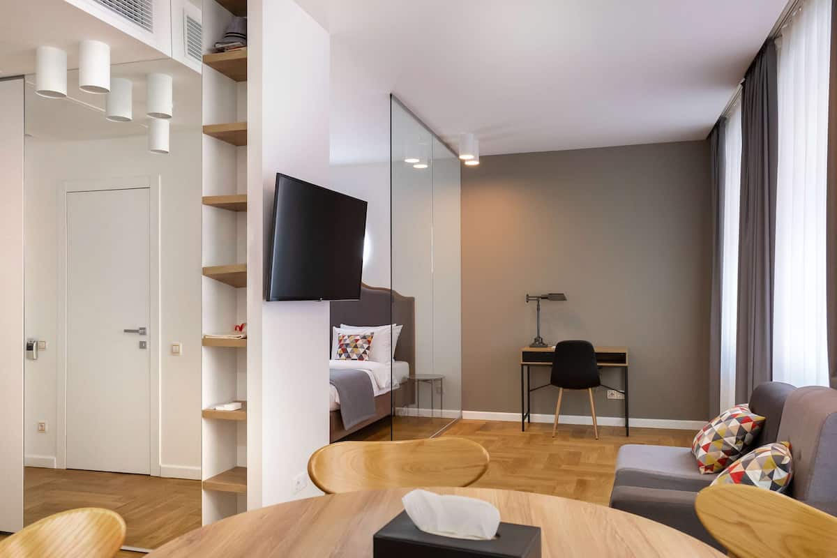 A modern hotel room with a bed, a desk, a TV, and a dining area with a table and chairs.