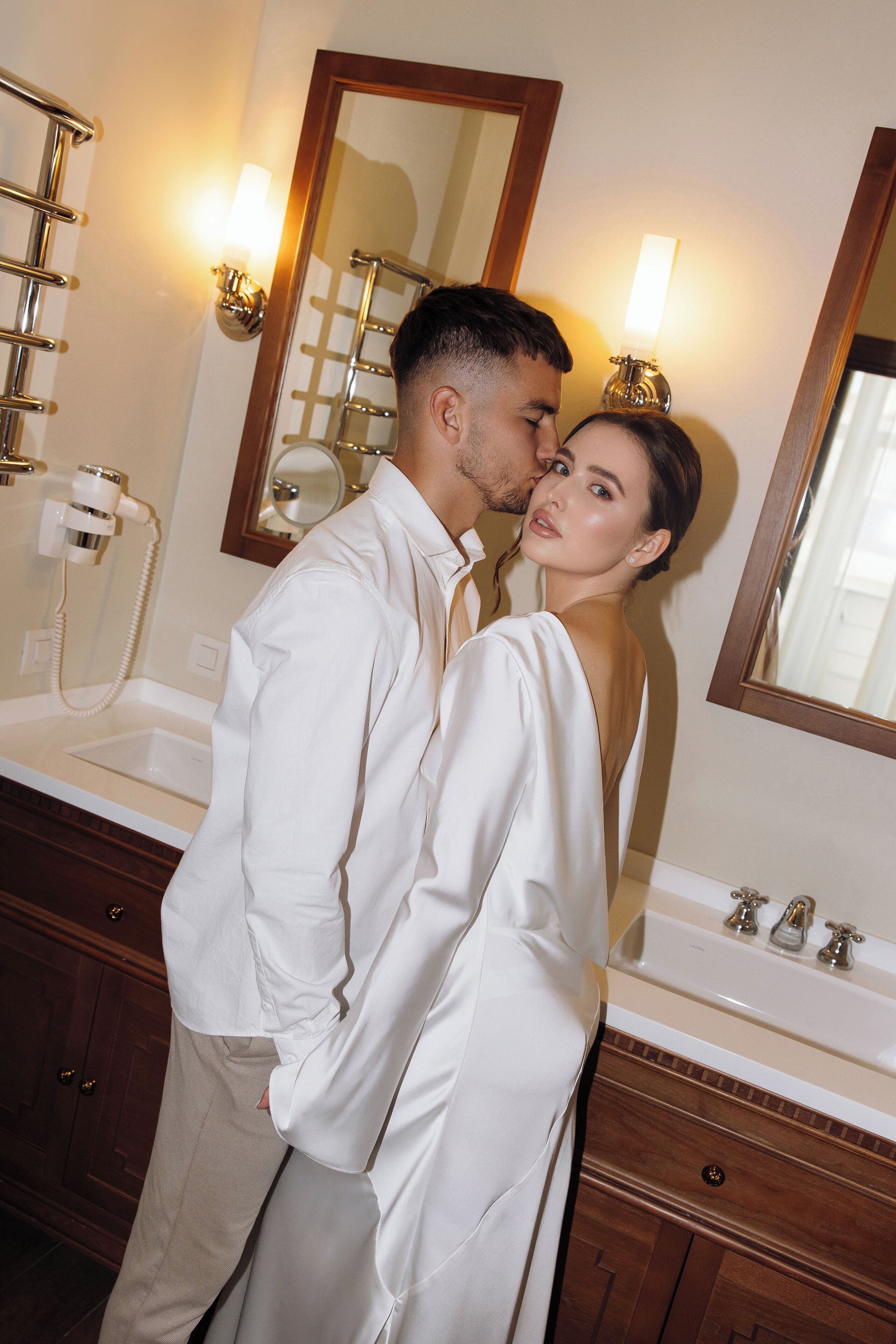 A man and woman standing in a bathroom with a large mirror, a sink, and a towel rack.