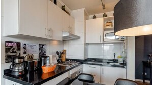 Moderni keittiö, jossa on valkoiset kaapit, mustat työtasot ja musta tuoli. Työtasolla on kahvinkeitin, vedenkeitin ja kori.