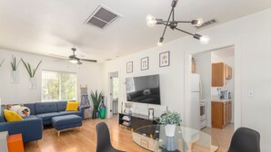 A modern living room with a blue sofa, a glass coffee table, and a TV. There is a kitchen area with wooden cabinets and a refrigerator. - Charming Bungalow In The Heart Of Downtown Phoenix (Phoenix)