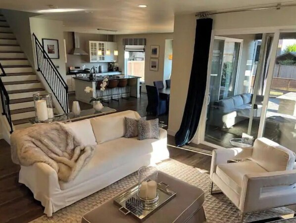 A modern living room with a sofa, armchair, and coffee table. A kitchen is visible in the background, and a staircase leads to another level.