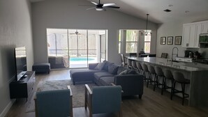 A spacious living room with a large sectional sofa, a dining area with bar stools, and a kitchen with white cabinets. - Cheerful 3 bedroom home with pool -- 5 min from beach! (Nokomis)