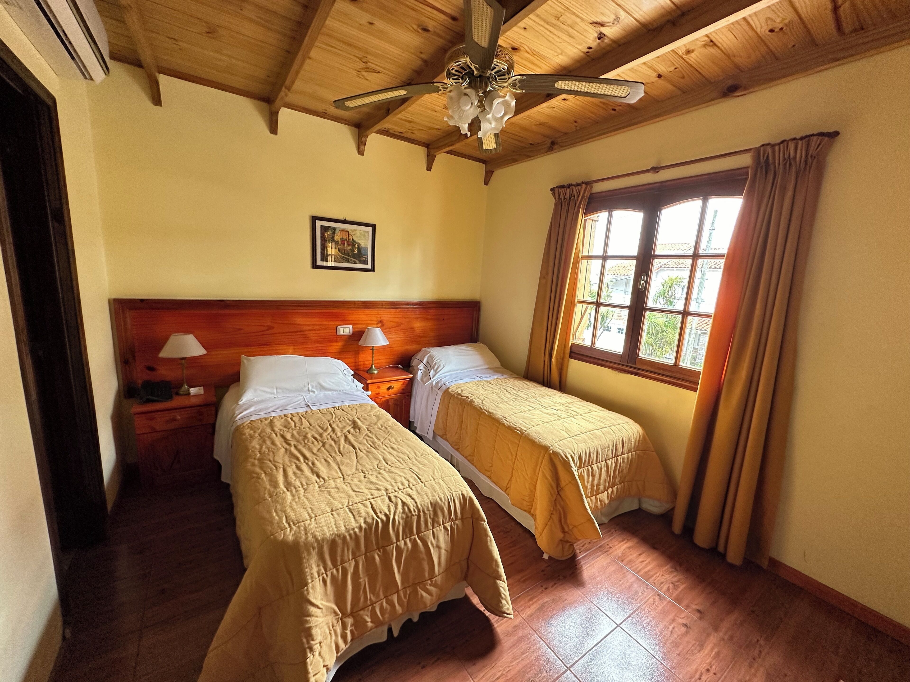 A room with two beds, wooden ceiling beams, a ceiling fan, a window with curtains, and a painting on the wall.