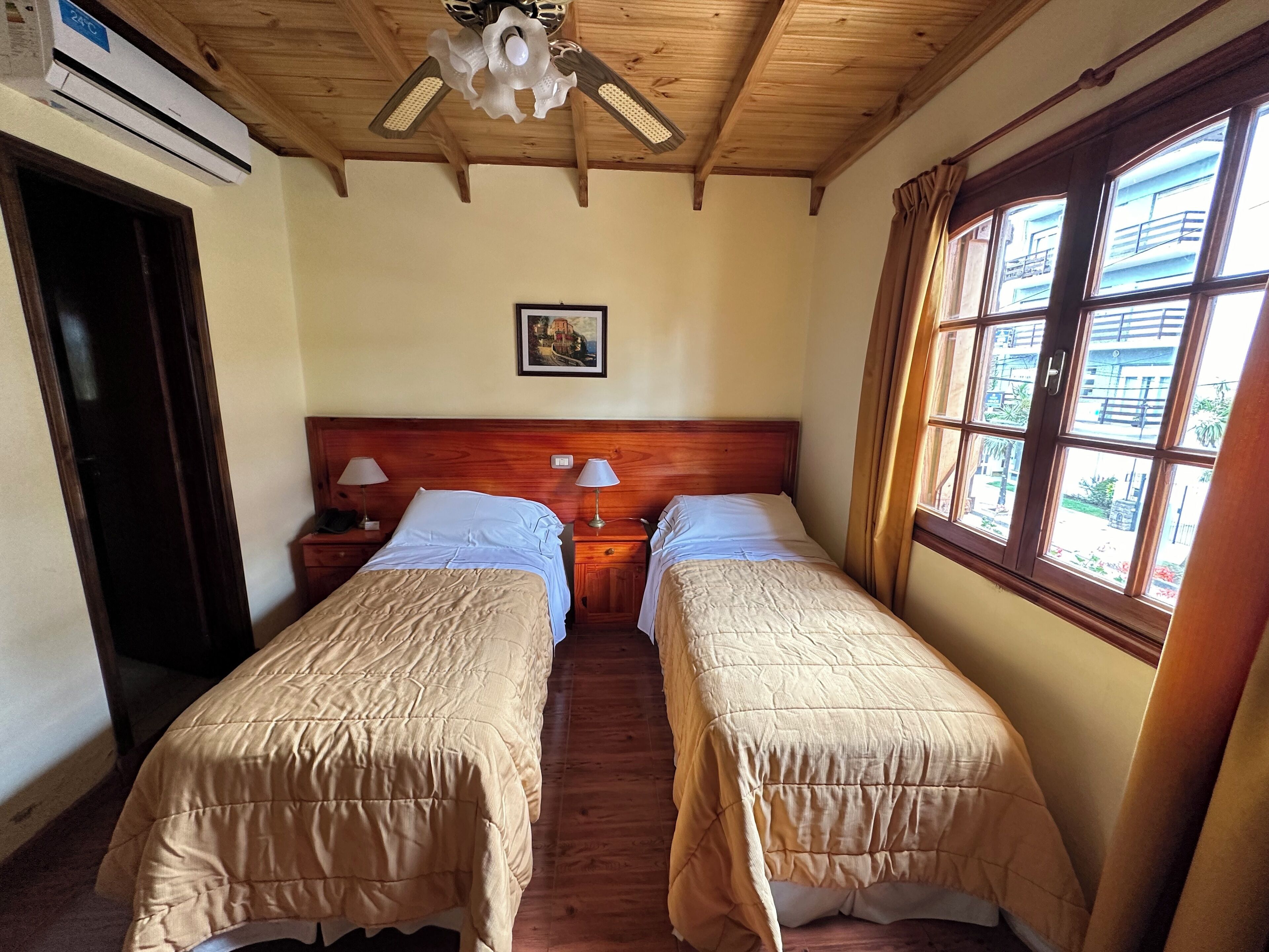A room with two beds, wooden headboards, a ceiling fan, and a window with a view of buildings.
