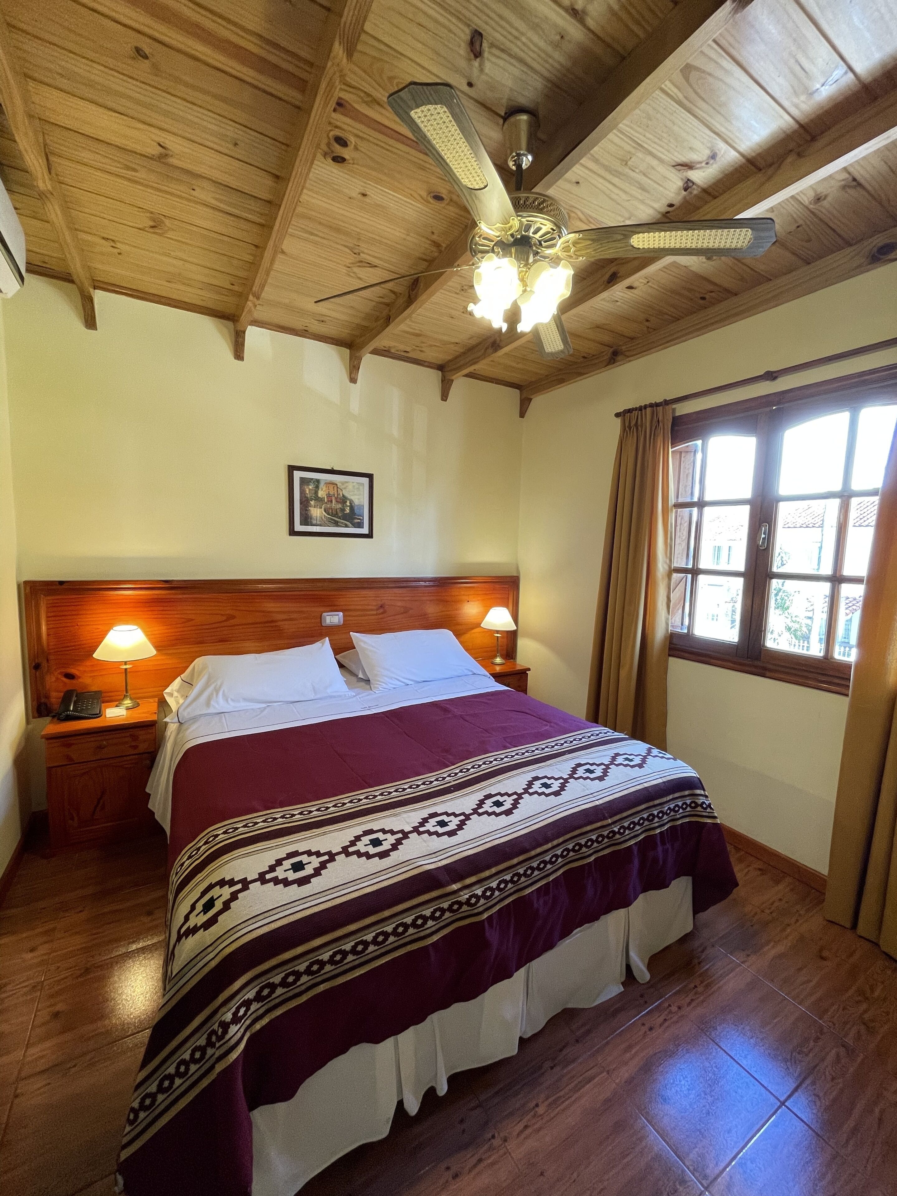 Un dormitorio con techo de madera, una cama con colcha estampada, dos lámparas de noche, una mesita de noche con un teléfono y una ventana con cortinas.