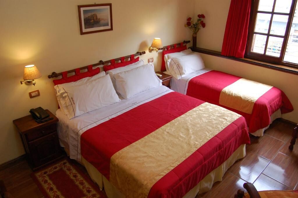 A hotel room with two beds, each with red and white bedding, a nightstand with a lamp, a wooden chair, and a window with red curtains.