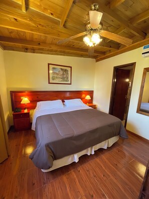 A bedroom with a wooden ceiling, a bed with a grey bedspread, two bedside lamps, a nightstand, a wooden door, and a framed picture on the wall.