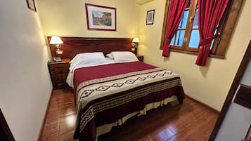 Une chambre à coucher avec une tête de lit en bois, un lit recouvert d’une couvre-lit à motifs, deux lampes de chevet, une fenêtre avec des rideaux rouges et un tableau encadré accroché au mur.