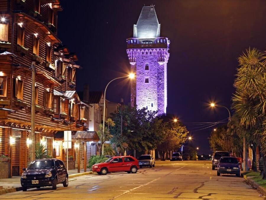 Eine Straße mit einem in Lila beleuchteten Turm, parkenden Autos und Häusern mit Balkonen.