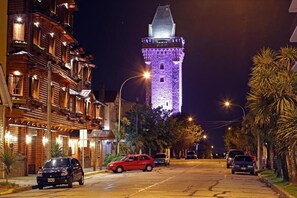A street with a tower illuminated in purple, parked cars, and buildings with balconies.