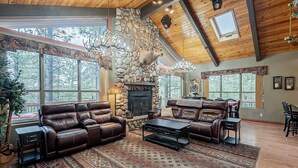 A spacious living room with a stone fireplace, leather sofas, and large windows offering a view of the forest. - Custom 6 Bedroom 4 full bathrooms Cabin with Amazing hillside Views, fenced lot. (Pinetop-Lakeside)