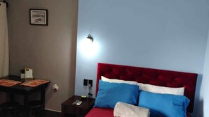 A bedroom with a red upholstered headboard, a bedside table with a lamp, and a small desk with a chair.