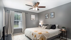 A bedroom with a bed, a ceiling fan, a window with curtains, a bedside table with a lamp, and artwork on the wall. - Wild Joy| King Bed, Pet-Friendly Stay, Fenced Yard (Clarksville)