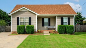A single-story house with a brick roof, beige siding, and black shutters. There is a front porch with a bench, and the property is surrounded by a well-maintained lawn and a wooden fence. - Wild Joy| King Bed, Pet-Friendly Stay, Fenced Yard (Clarksville)
