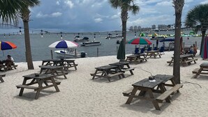 A beach with wooden picnic tables and umbrellas, palm trees, and boats in the background. - New! GREEN HOUSE minutes from Navarre beach and restaurants (Navarre)