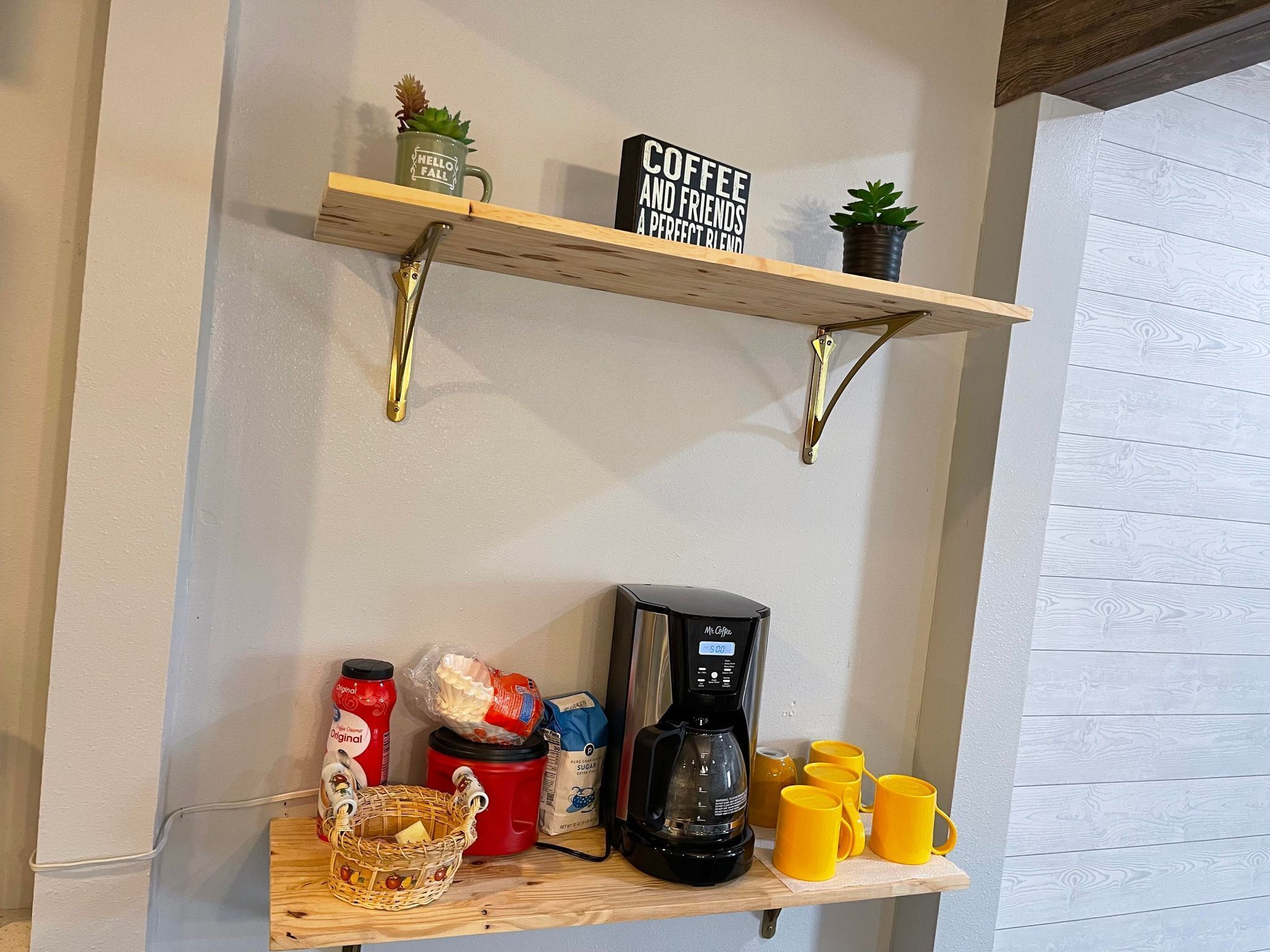 En kökshylla med en kaffebryggare, gula muggar och en korg med frukt.