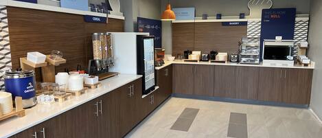 A modern breakfast area with a self-serve station, including a refrigerator, coffee machine, and various food items.