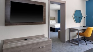 A hotel room with a large flat-screen TV mounted on the wall, a wooden dresser with drawers, a desk with a chair, and a bed in the background. - Holiday Inn Express Rocky Mount Sports Center by IHG (Rocky Mount)