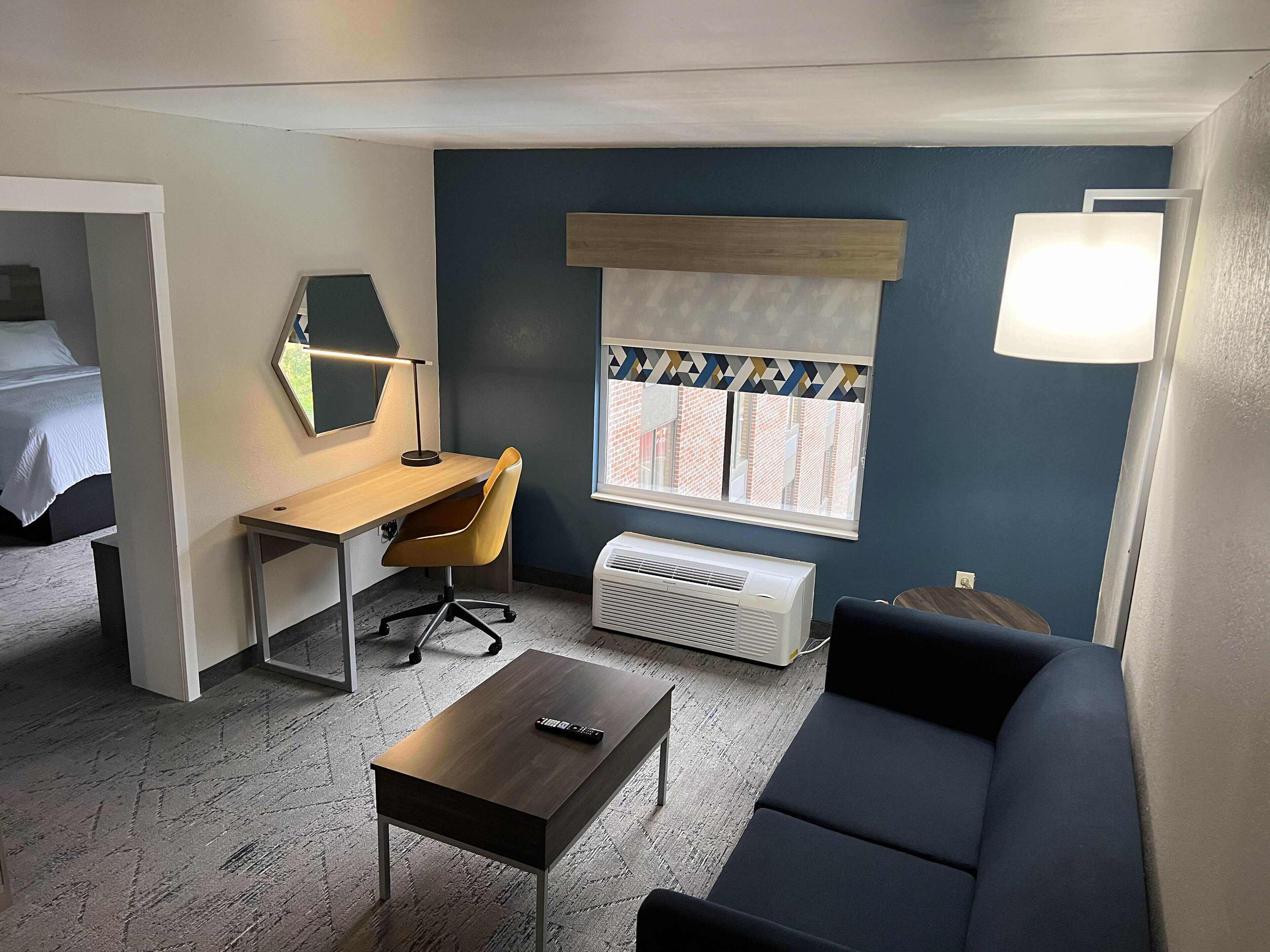 Habitación de hotel compacta con cama, escritorio con silla, una mesita, un sofá, ventana con persianas y una lámpara de pared.