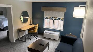 Habitación de hotel compacta con cama, escritorio con silla, una mesita, un sofá, ventana con persianas y una lámpara de pared.