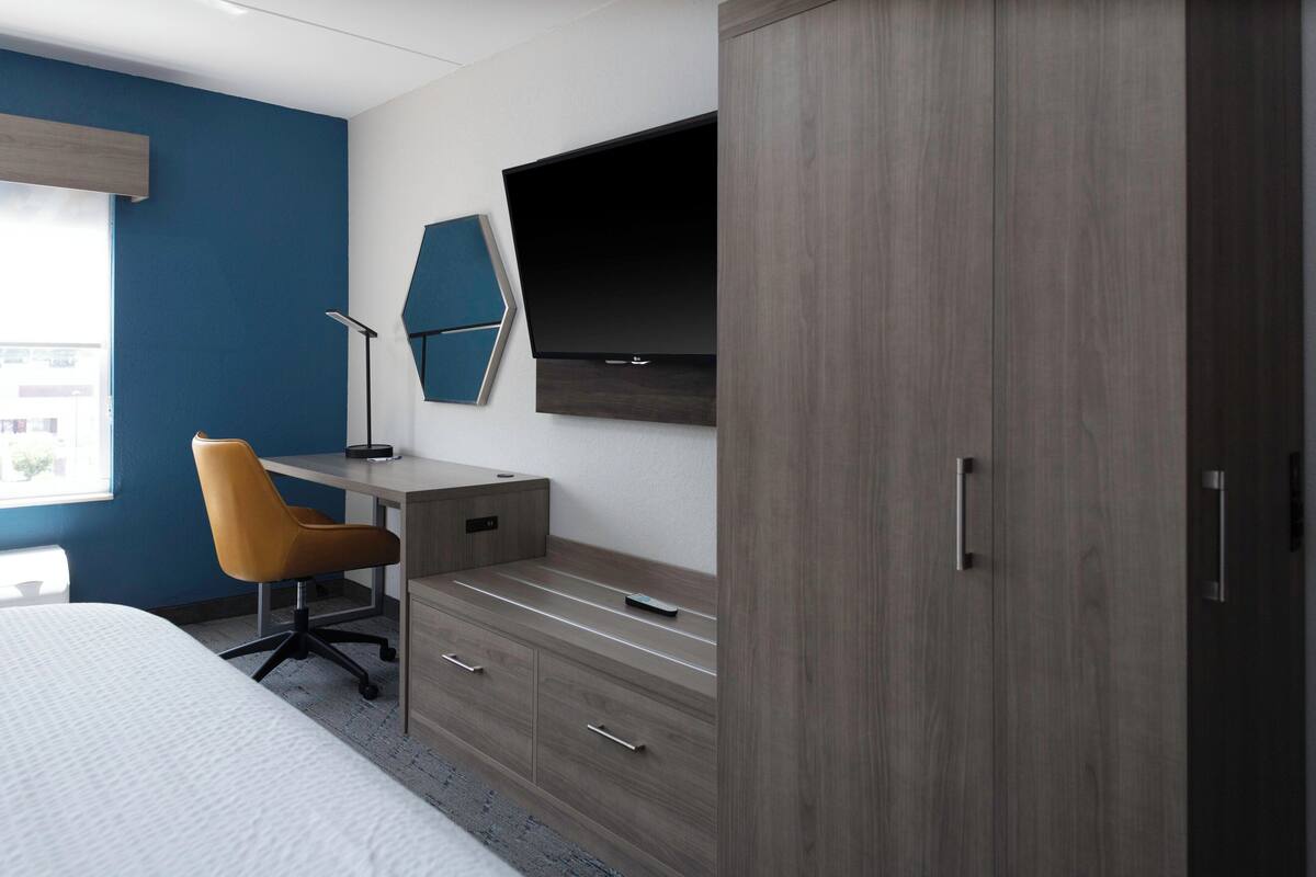 A modern hotel room with a bed, a desk with a chair, a TV mounted on the wall, and a wardrobe.