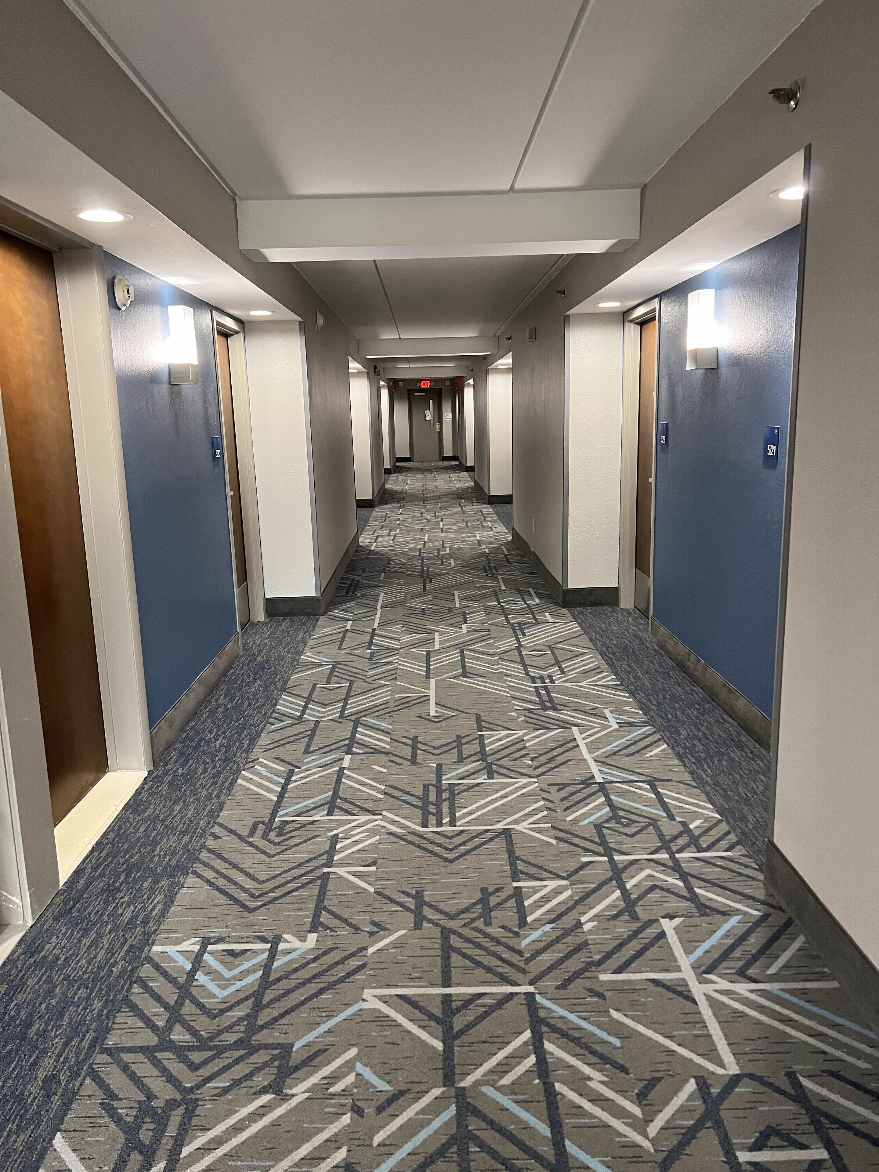 Un pasillo de hotel con alfombra estampada, puertas a ambos lados y techo con iluminación empotrada.