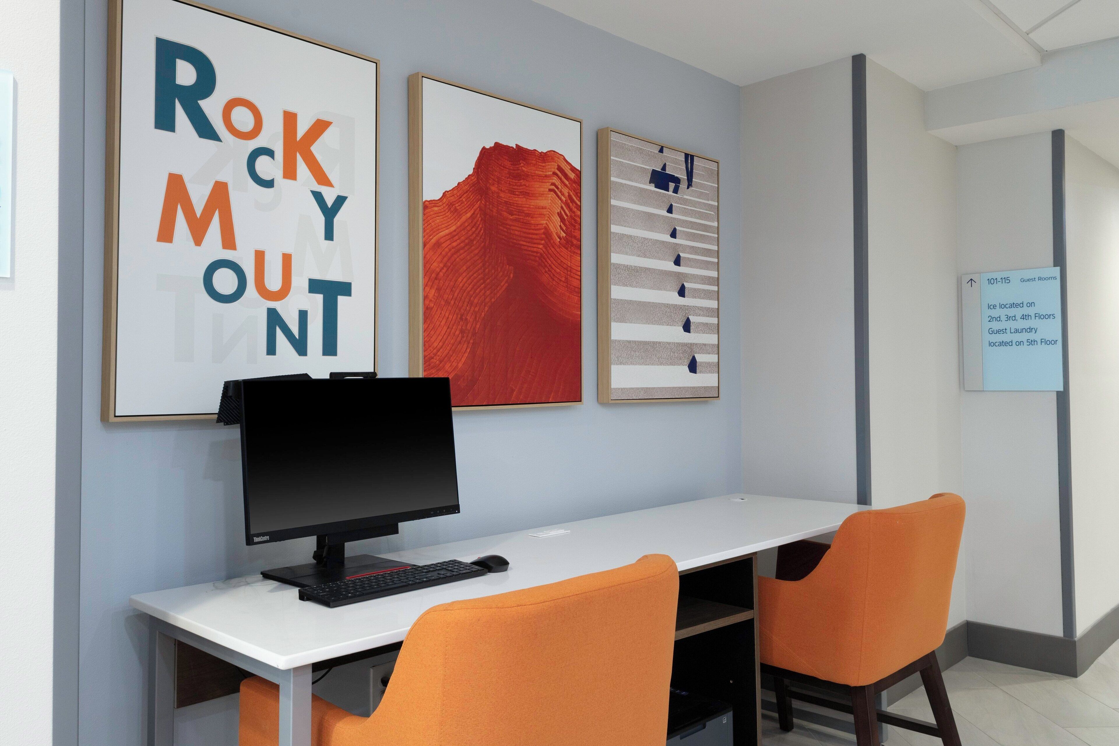 Una habitación de hotel con un escritorio, dos sillas naranjas, una computadora y tres obras de arte enmarcadas en la pared.