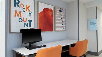 Una habitación de hotel con un escritorio, dos sillas naranjas, una computadora y tres obras de arte enmarcadas en la pared.