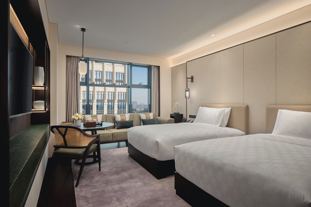Habitación de hotel con dos camas, un sofá, una mesa de comedor y vistas a un paisaje urbano a través de amplias ventanas.