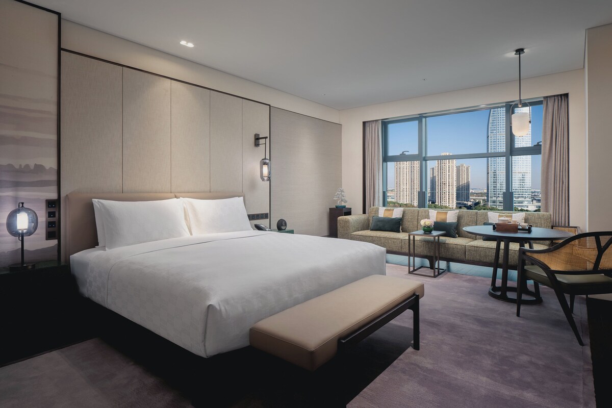 Habitación de hotel moderna con una cama grande, un sofá, un comedor y vistas a edificios altos.