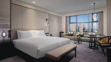 A modern hotel room with a large bed, a sofa, a dining area, and a view of tall buildings.