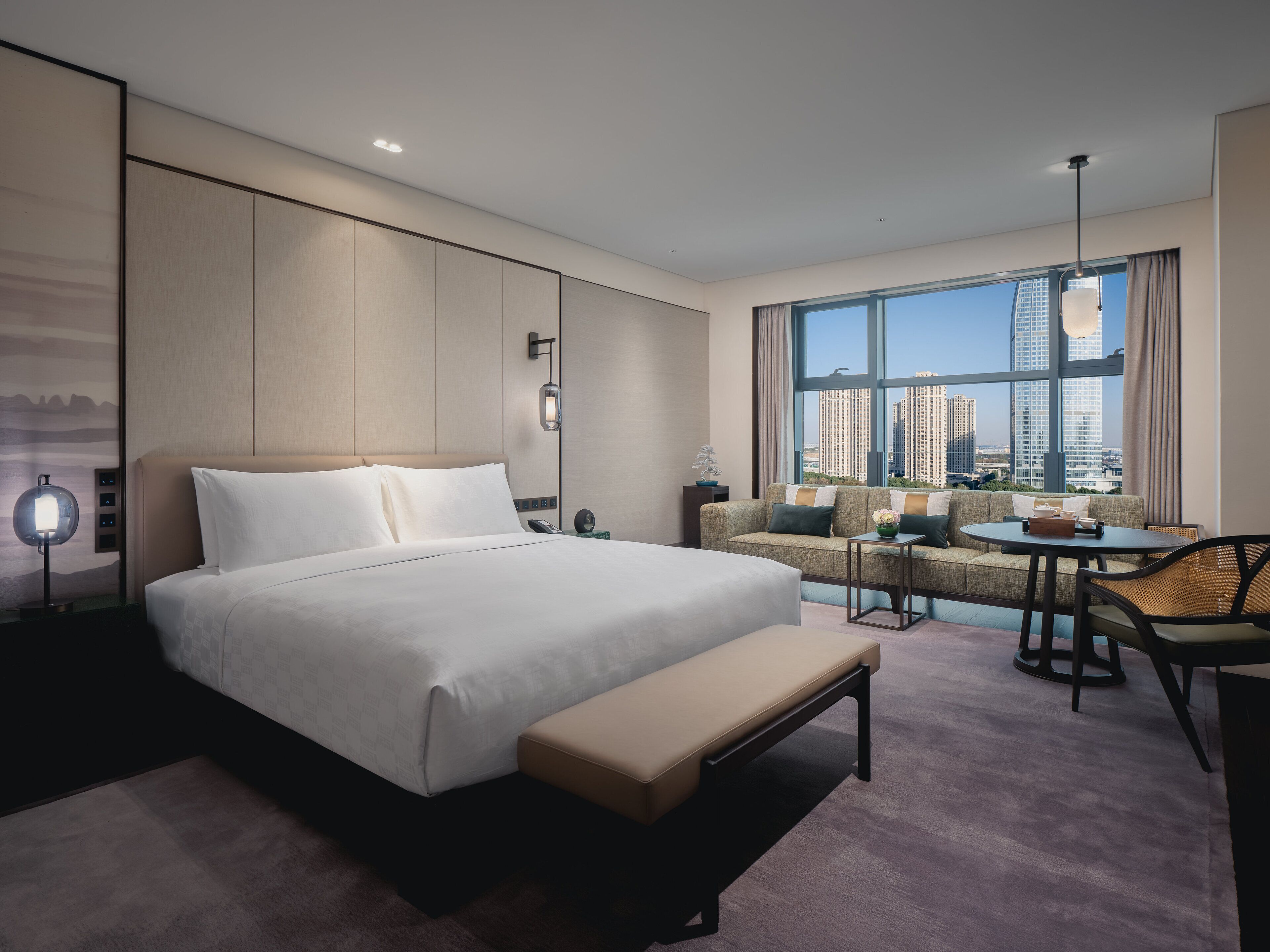 Ein modernes Hotelzimmer mit einem großen Bett, einer Sitzecke, einem Essbereich und Blick auf hohe Gebäude.