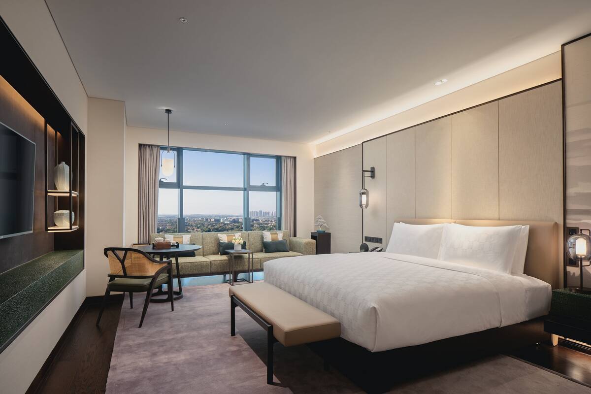 Camera d'albergo moderna con un letto grande, una zona relax con un tavolo e vista sulla città attraverso ampie finestre.