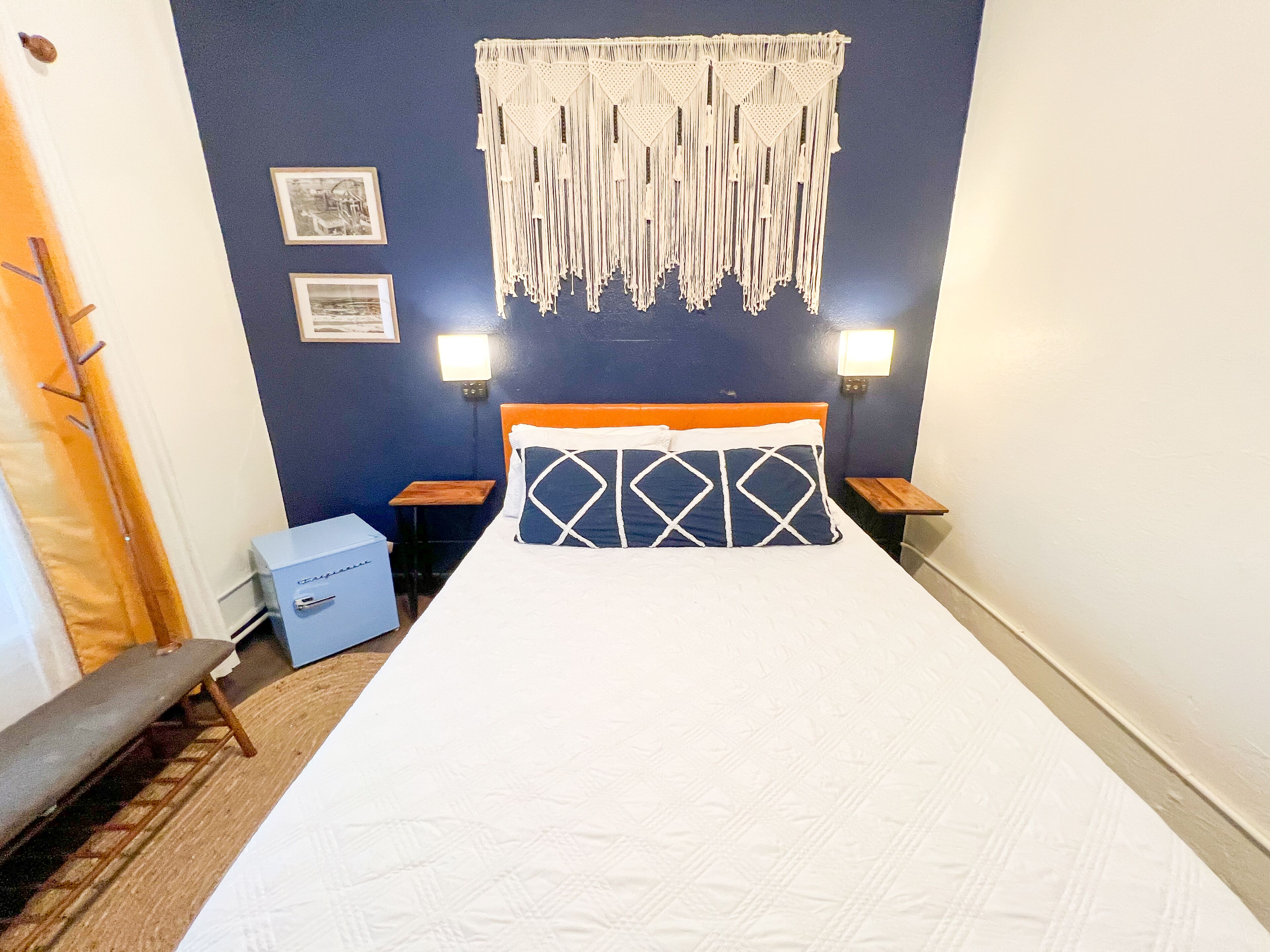 Un lit bien fait, agrémenté d’un coussin à motifs, d’une décoration murale en macramé et de deux lampes de chevet.