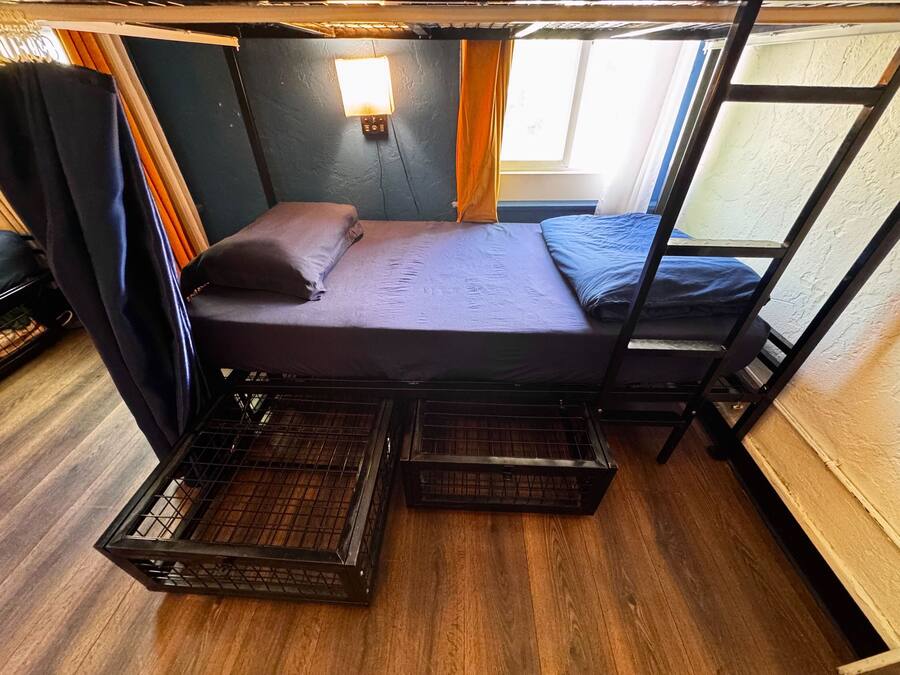 Uma cama beliche com colchão, travesseiros e cabeceira. Há uma janela com cortinas e uma luminária fixada na parede.