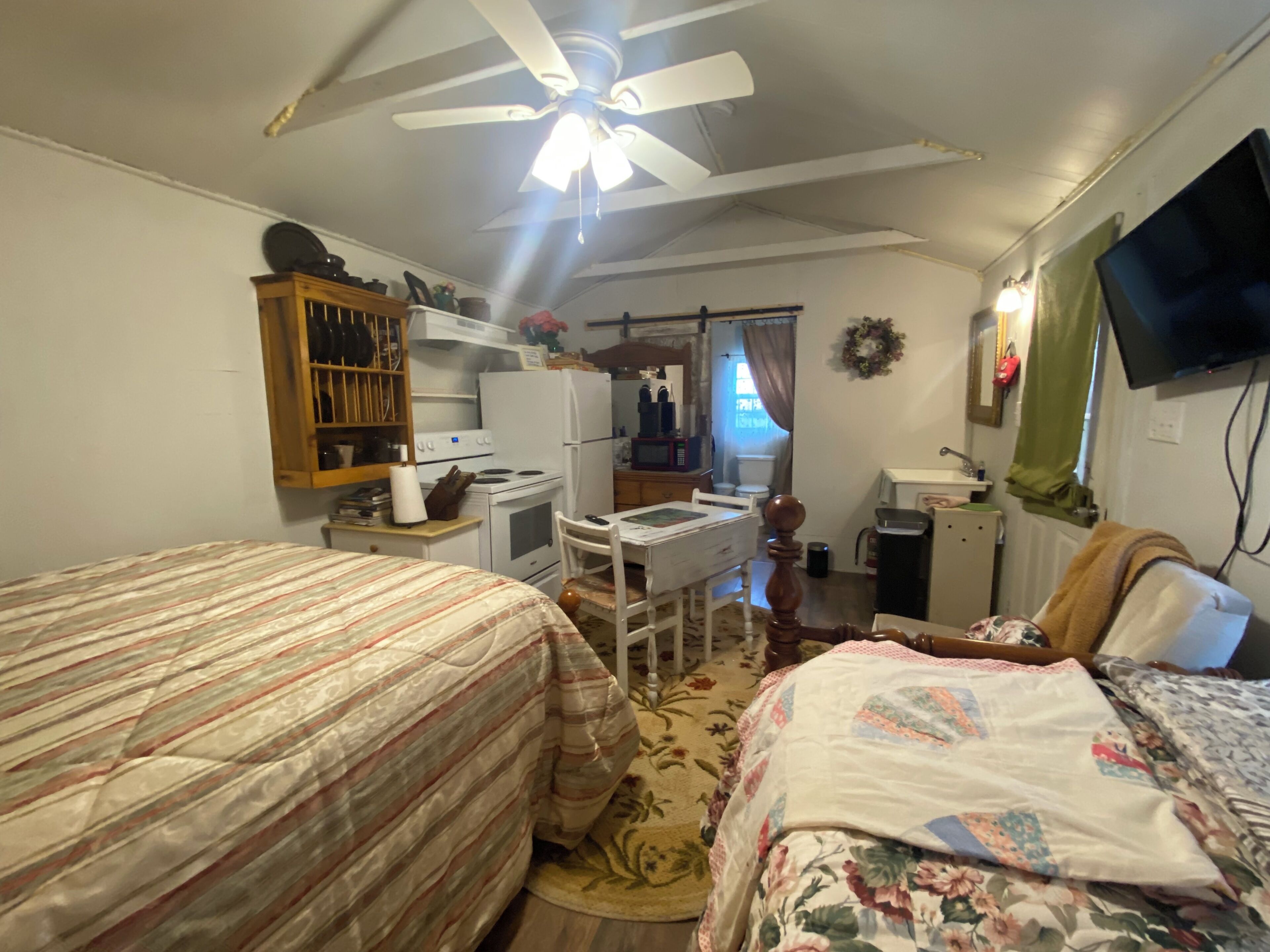 A bedroom with two beds, a desk, a chair, a ceiling fan, a television, and a kitchenette.