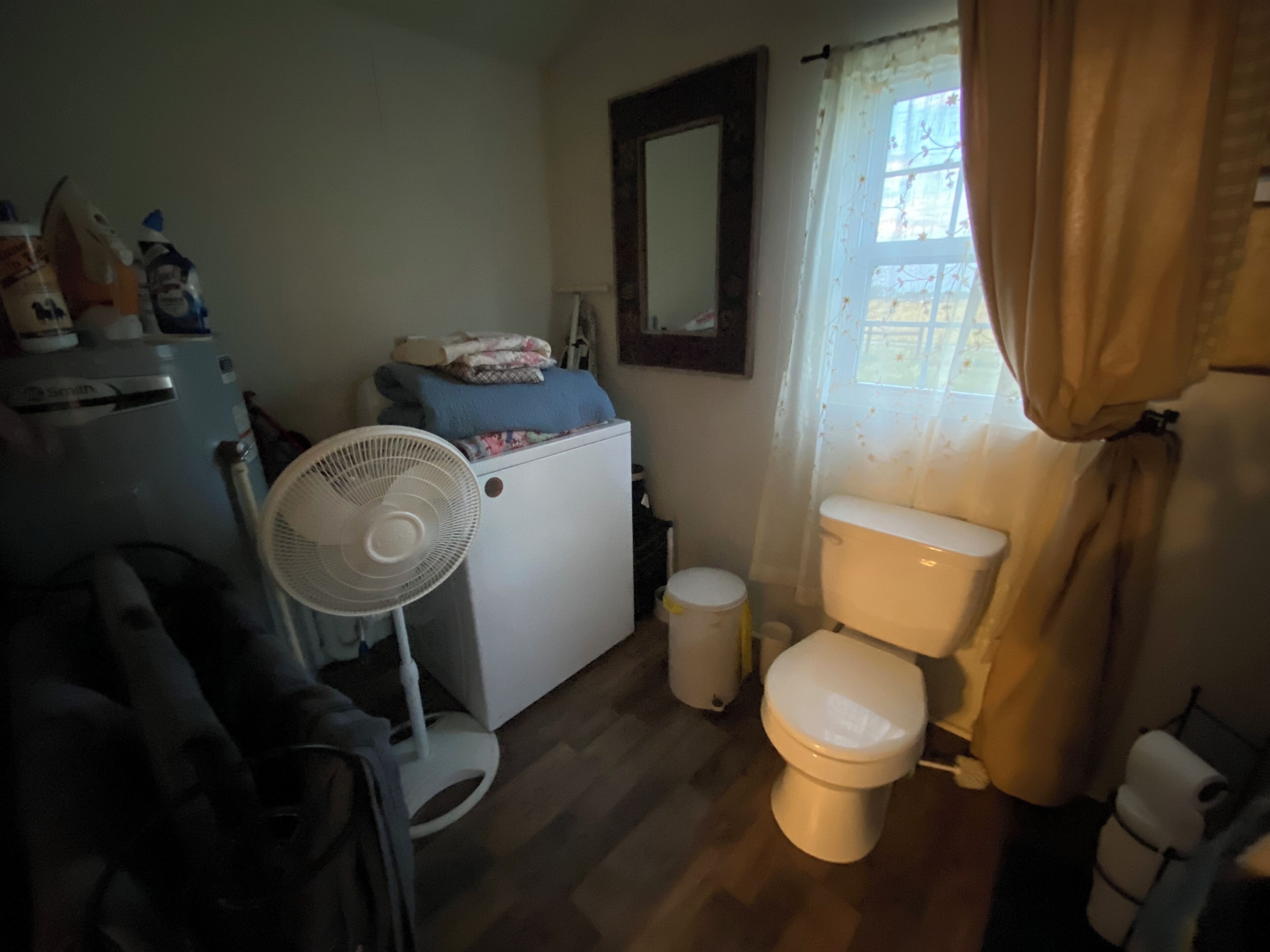 Une petite pièce équipée d’un lave-linge, d’un ventilateur, de toilettes et d’une petite fenêtre avec des rideaux.