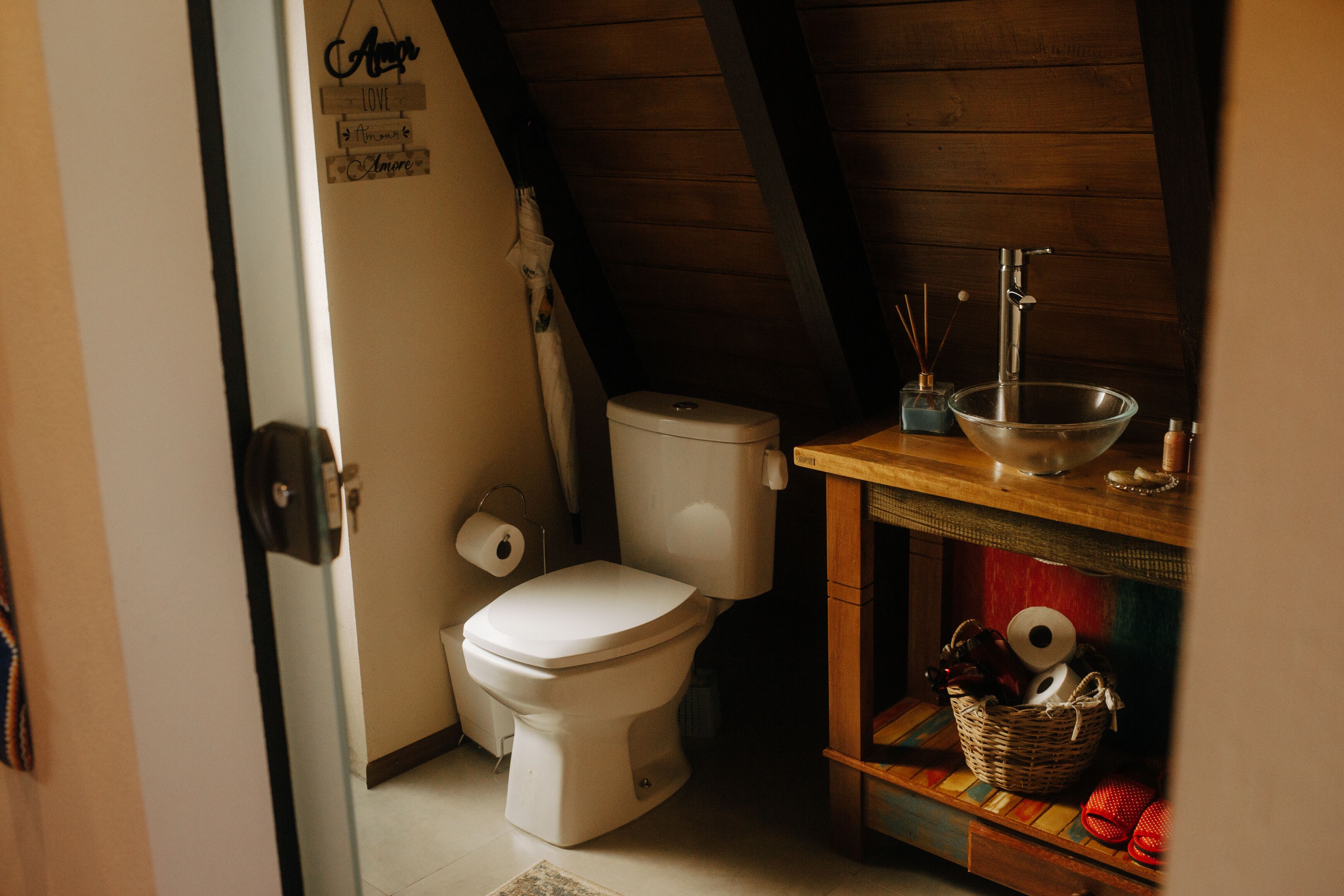 A small, rustic bathroom with a wooden ceiling, a white toilet, a wooden shelf with a bowl and toiletries, and a wall with a 'Love' sign.