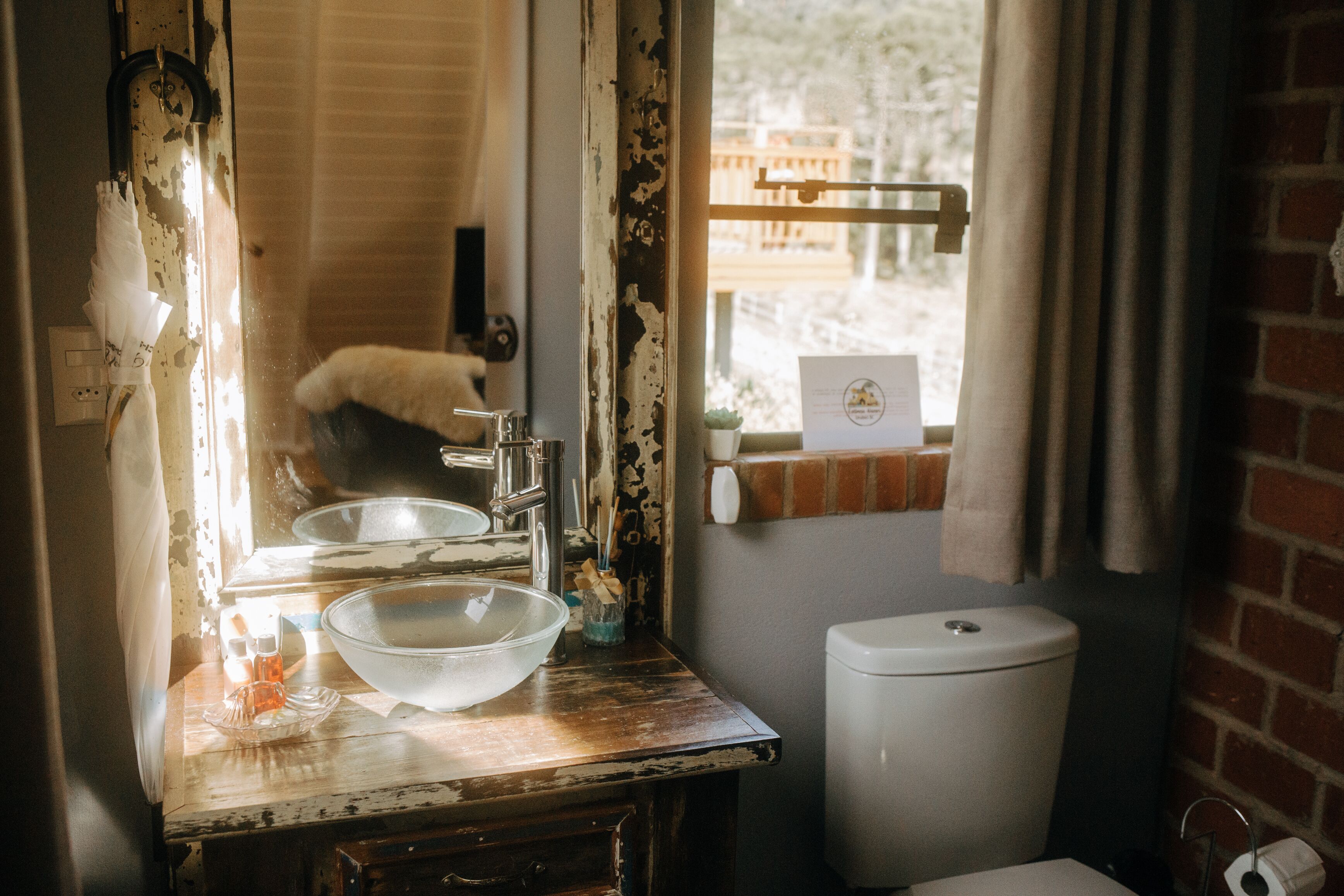 Un baño rústico con tocador de madera, un lavabo grande de vidrio, inodoro blanco y ventana con vista a una terraza de madera.