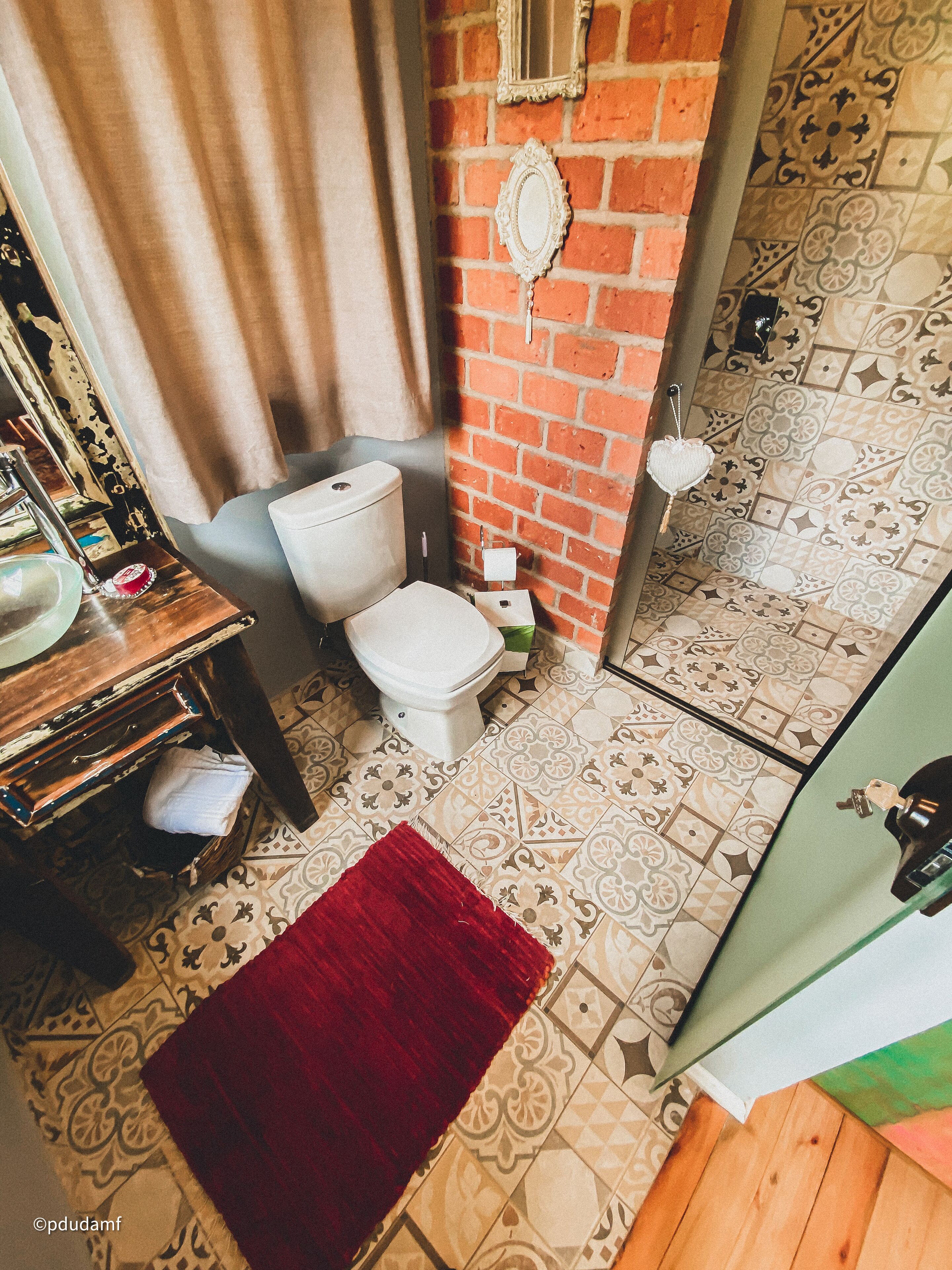 Ein Badezimmer mit Toilette, Waschbecken und Spiegel an einer Ziegelwand. Der Boden ist mit einem Muster versehen und ein roter Teppich liegt darauf.
