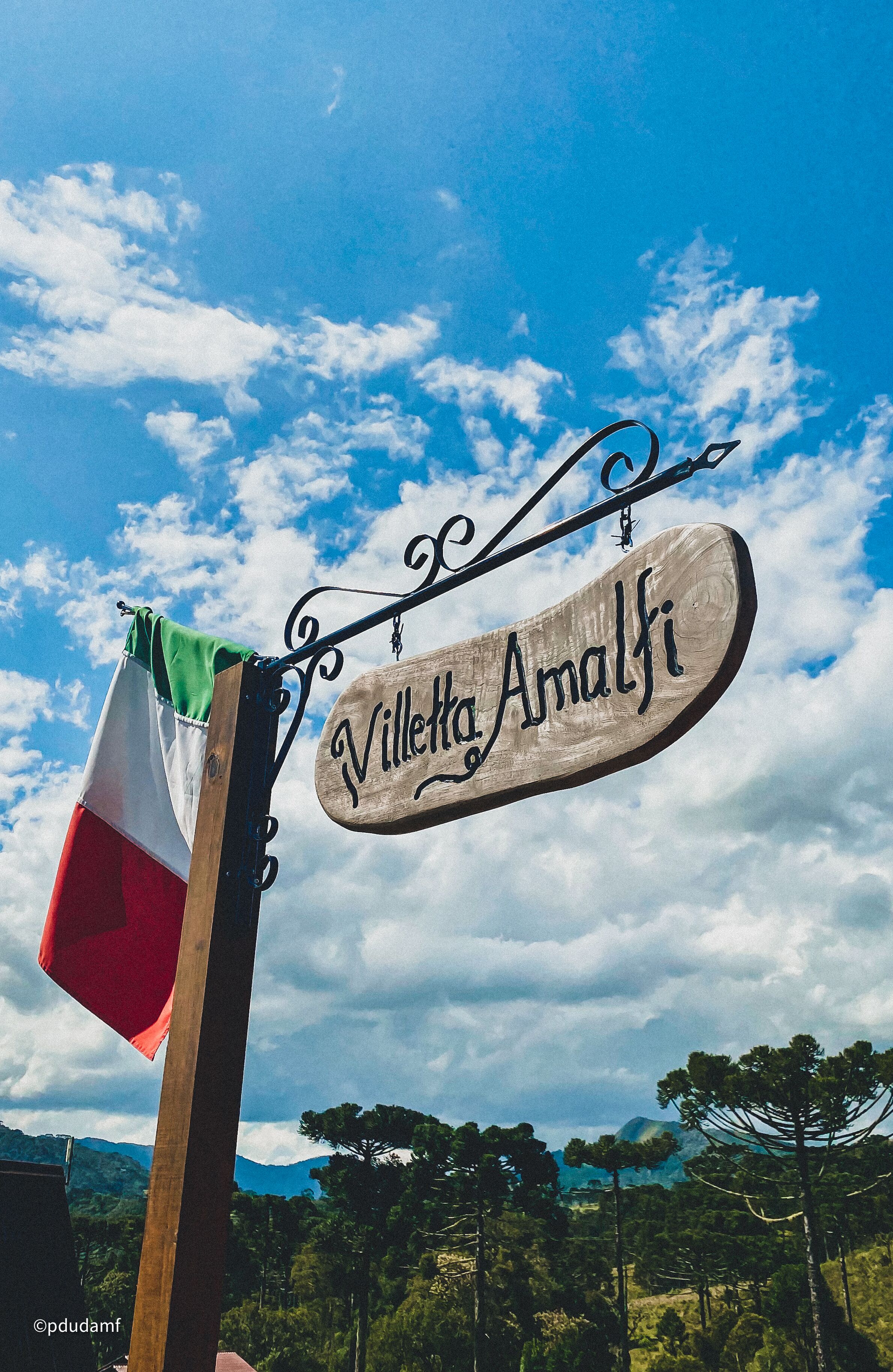 Een houten bord met de naam "Villette Amalfi" voor een blauwe lucht met wolken en een vlag.