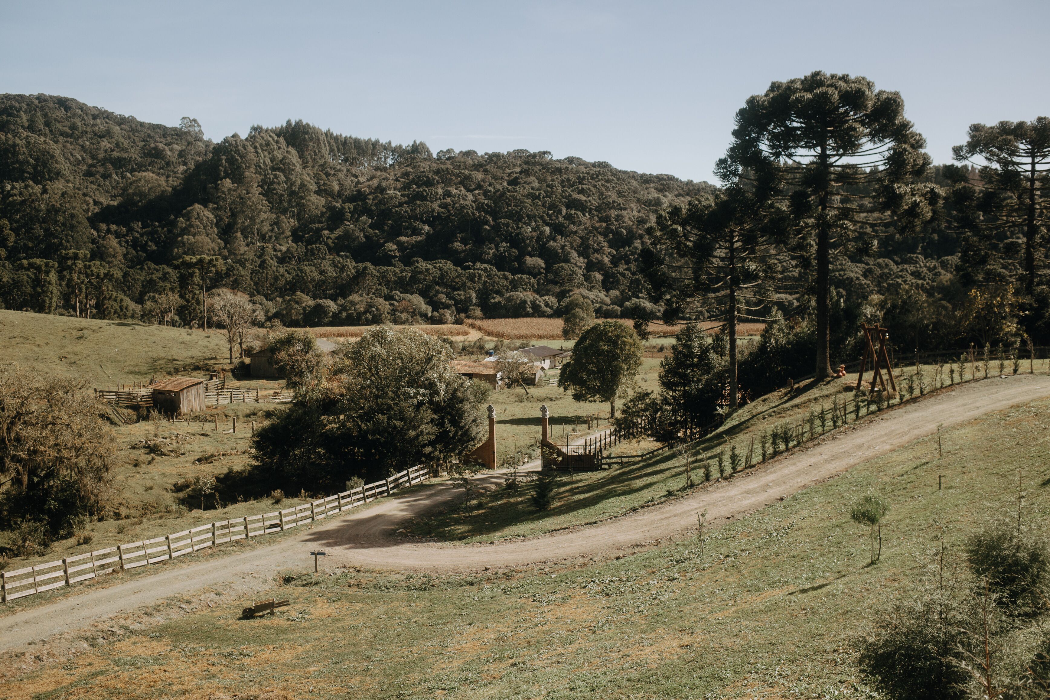 Paisaje rural con camino de tierra, cerca de madera y edificios dispersos.