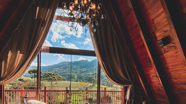 A cozy room with a wooden ceiling, a large window with curtains, a wooden deck outside, and a hot tub. - Estância Nazari (Urubici)