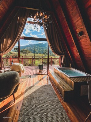 A cozy room with a wooden ceiling, a large window with curtains, a wooden deck outside, and a hot tub. - Estância Nazari (Urubici)