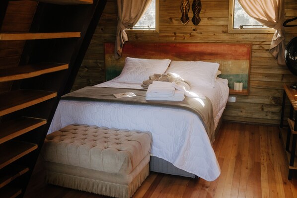 A cozy bedroom with a wooden headboard, a neatly made bed, a small ottoman, and a window with curtains. - Estância Nazari (Urubici)