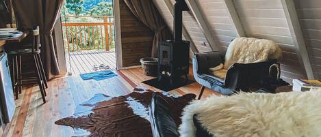 A cozy attic room with a sloped ceiling, a rustic wooden floor, a large cowhide rug, a fluffy white chair, and a black stove.